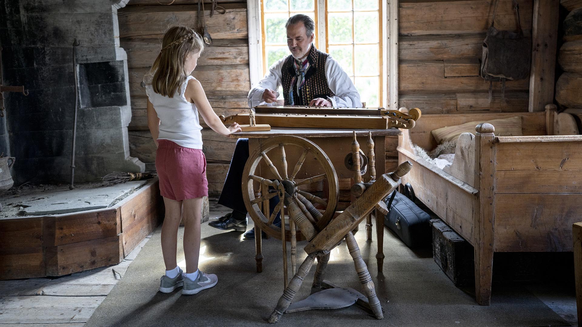 Inside one of the old houses at Valdres Folk Museum with a bed, a spinning wheel, a fire place and a man in folk costume who plays the langeleik, a kind of zither, while girl watches and listenes.