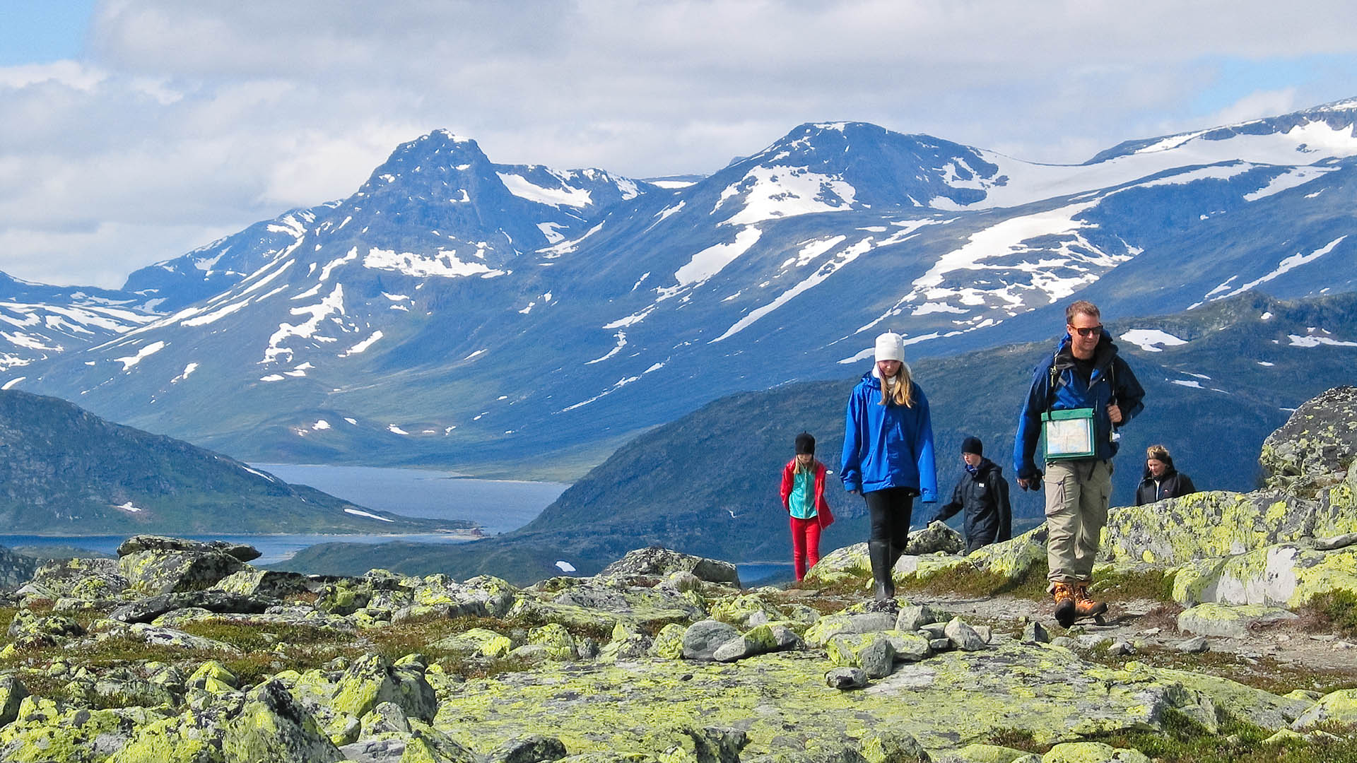 Hikers in the mountain pass a section with a lot of lichen-covered rocks. A lake and high mountains in the background.